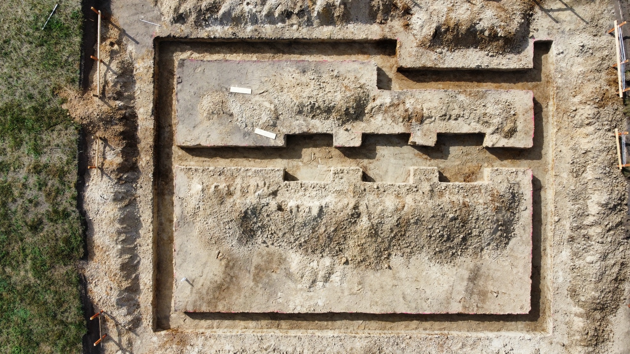 Widok z góry na wykop pod fundamenty domu jednorodzinnego z zaznaczonym obrysem ław fundamentowych, widoczne drewniane stemple i poziomica na trawie obok wykopu.