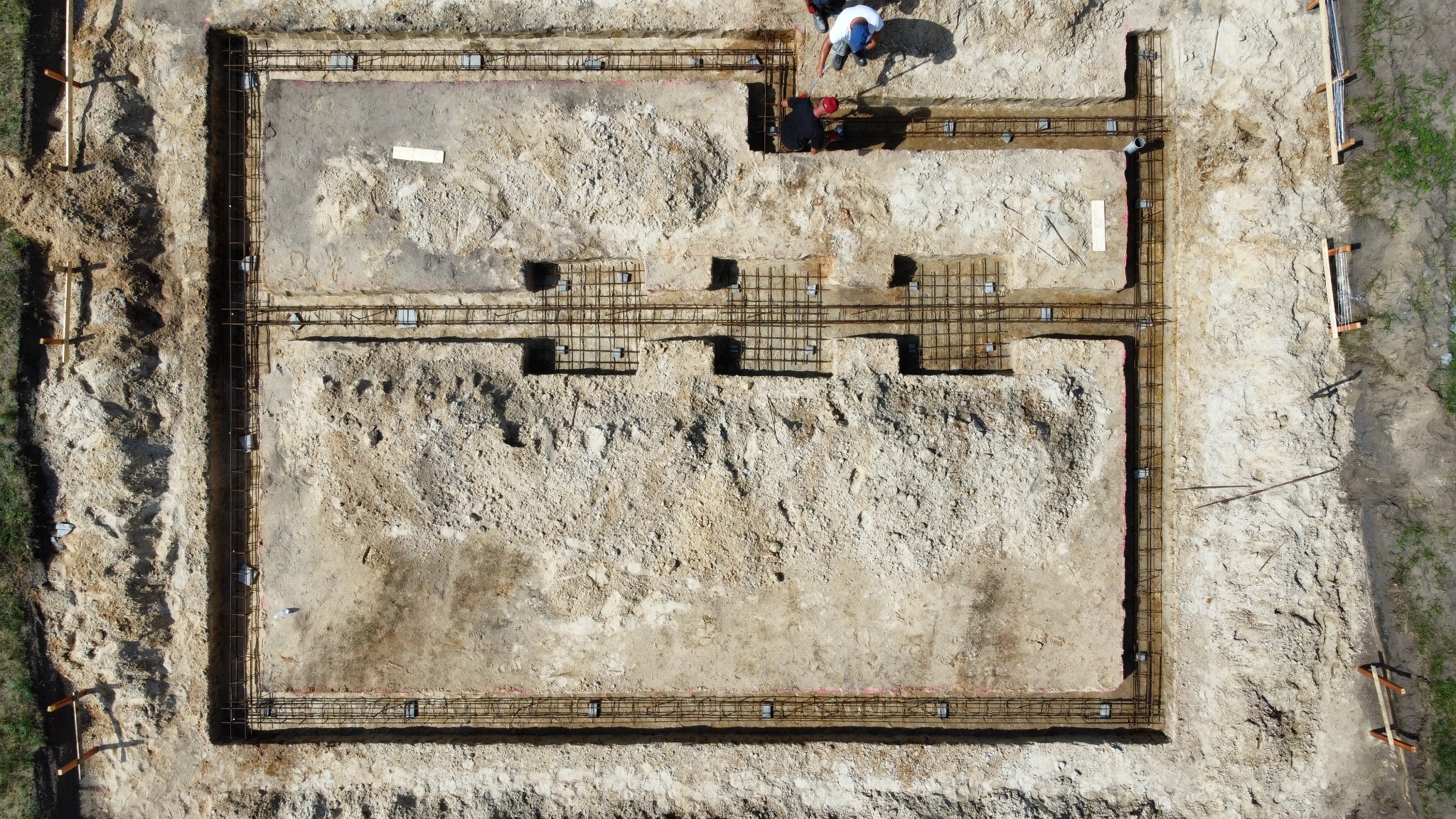 Widok z góry na wykop pod fundamenty z ułożonym zbrojeniem, dwóch robotników pracuje w rogu wykopu.