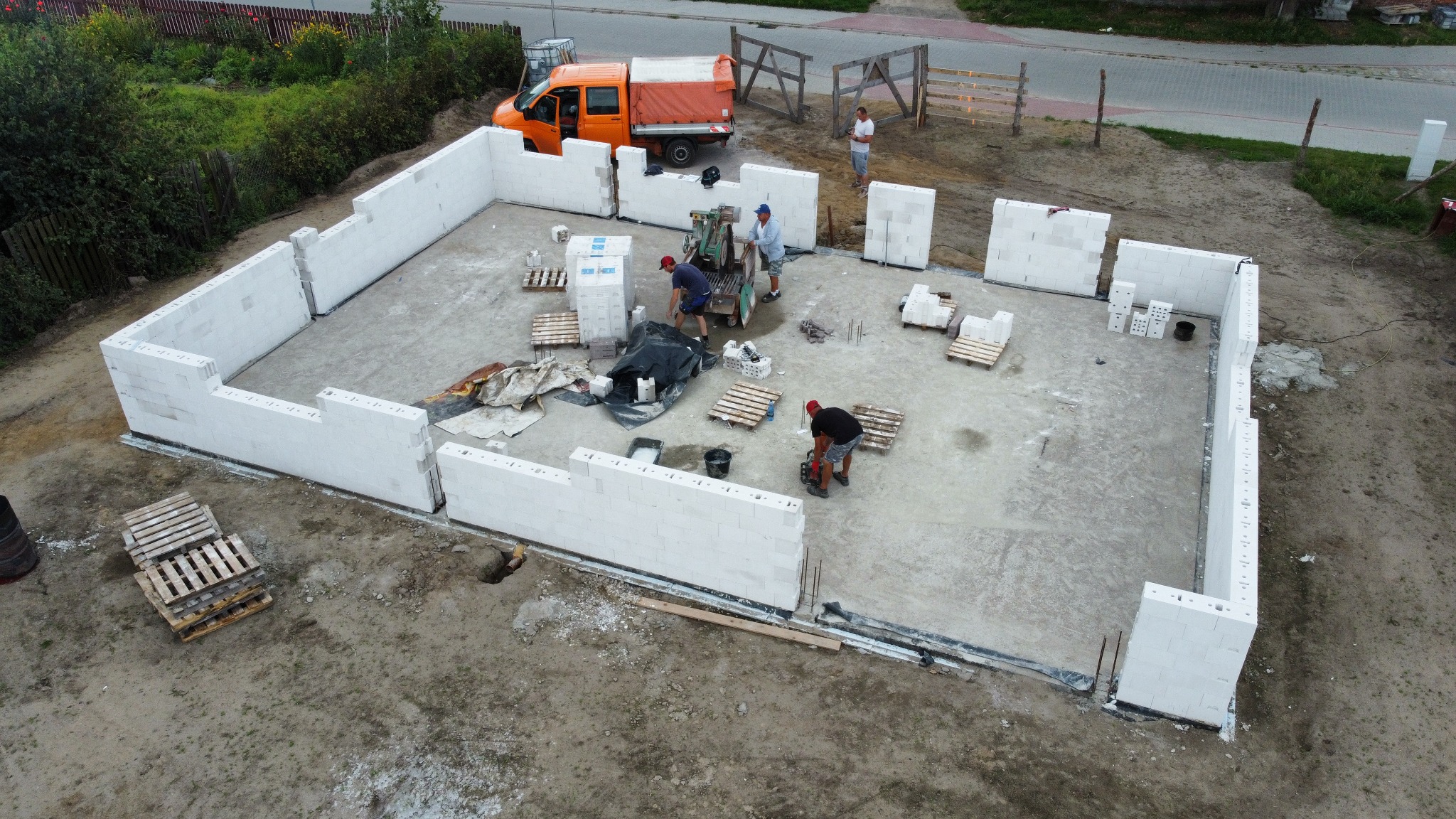 Budowa ścian fundamentowych z bloczków betonowych Ytong, widok z góry na plac budowy, pracownicy w trakcie murowania, pomarańczowy samochód dostawczy w tle.