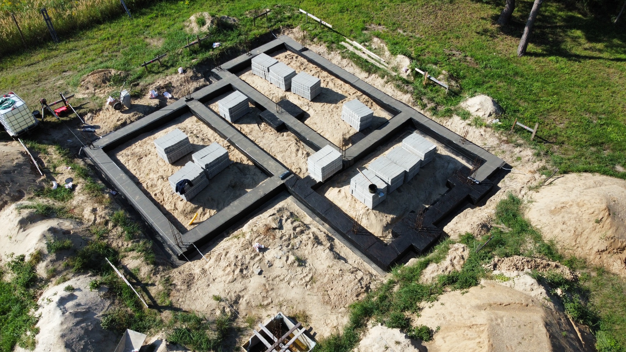 Widok z góry na wylane fundamenty budynku z izolacją przeciwwilgociową, częściowo obsypane piaskiem, z ułożonymi bloczkami betonowymi, w tle trawa i drzewa.