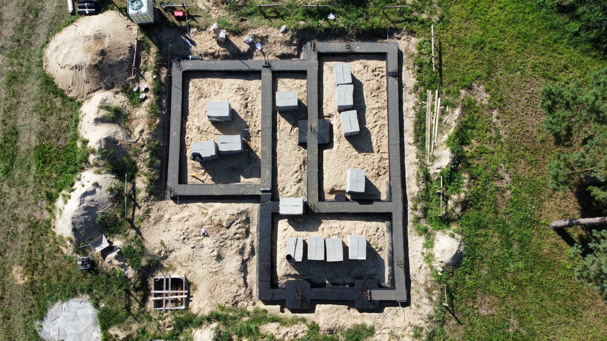 Widok z góry na zbrojone, czarne fundamenty domu w trakcie budowy, z ułożonymi bloczkami betonowymi wewnątrz wykopu, obok sterta piasku i sprzęt budowlany.