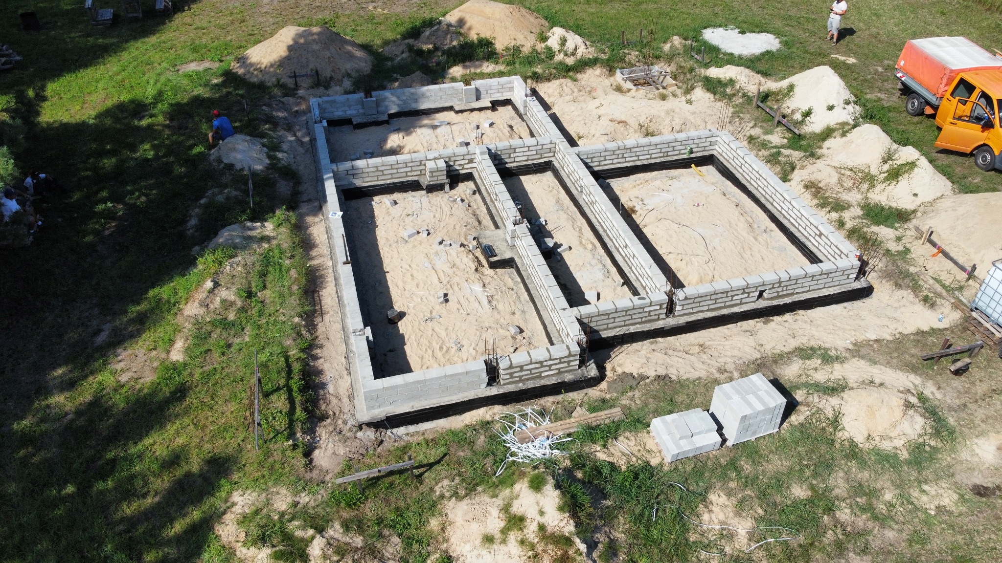 Widok z góry na wznoszone fundamenty budynku z bloczków betonowych na piaszczystym gruncie, izolacja przeciwwilgociowa widoczna, w tle hałda piasku i zaparkowany pomarańczowy samochód dostawczy.