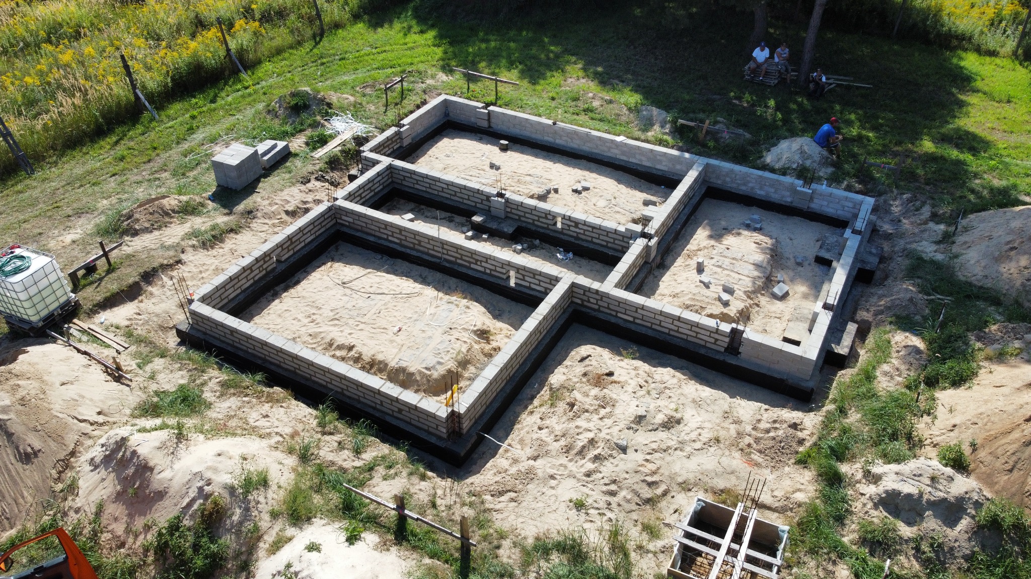 Widok z góry na fundamenty domu w trakcie budowy, widać bloczki betonowe tworzące ściany fundamentowe, piasek wewnątrz, teren budowy i osoby odpoczywające w tle.