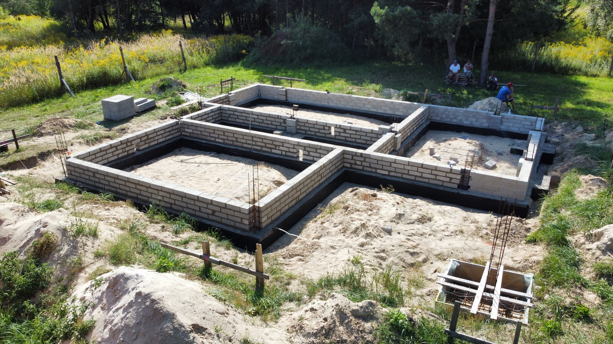 Budowa fundamentów domu jednorodzinnego: widok z góry na wzniesione ściany fundamentowe z bloczków betonowych, wypełnione piaskiem, z widoczną izolacją przeciwwilgociową, zbrojeniami i pracownikami...