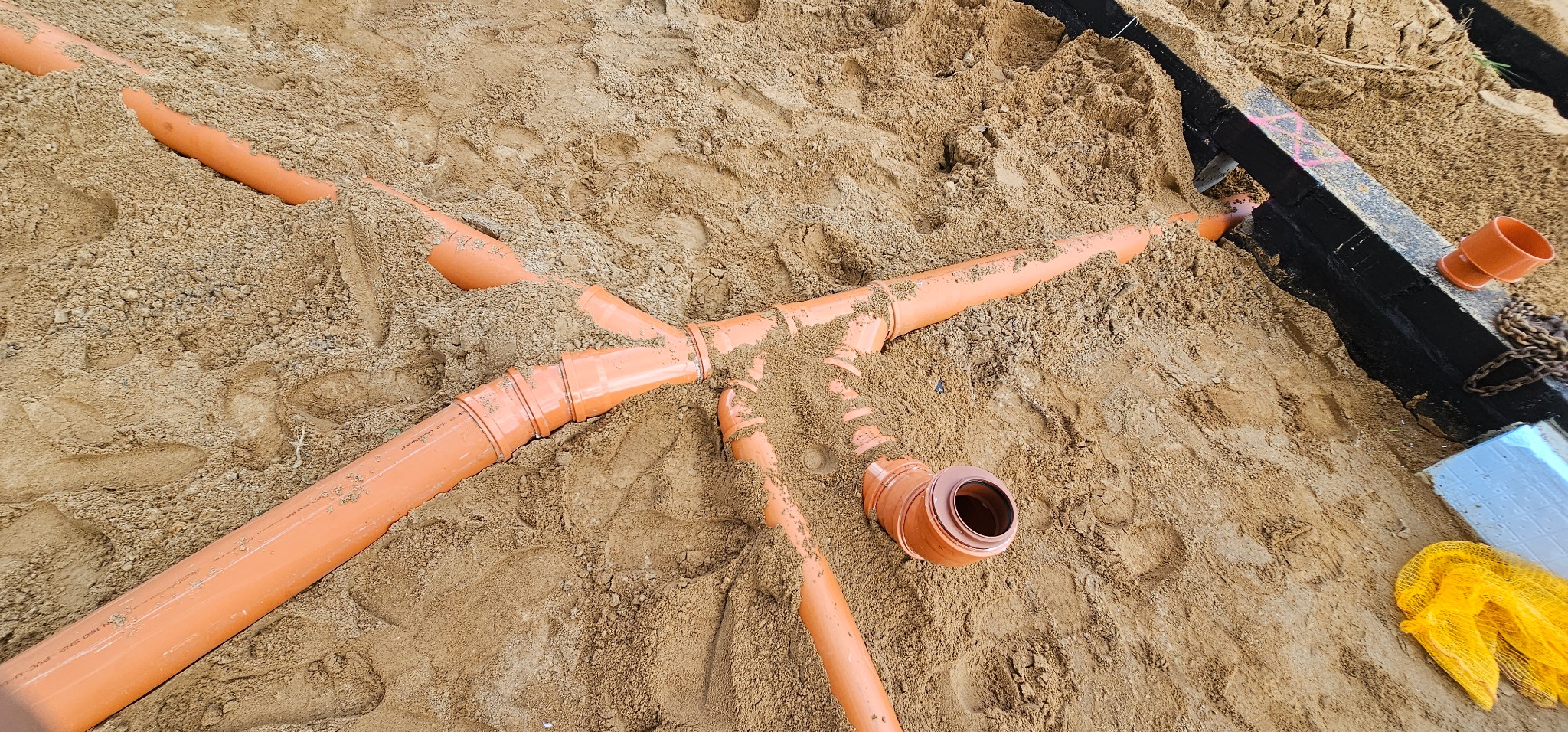 Pomarańczowe rury kanalizacyjne ułożone w piasku podczas budowy fundamentów, widoczne złącza i kształtki.