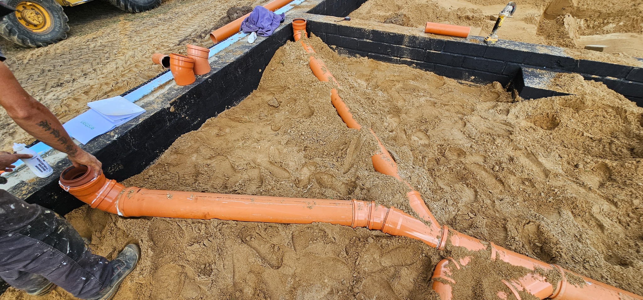 Układanie pomarańczowych rur kanalizacyjnych w wykopie fundamentowym, częściowo zasypanym piaskiem, z widocznymi izolowanymi ścianami fundamentów i pracownikiem łączącym elementy systemu.