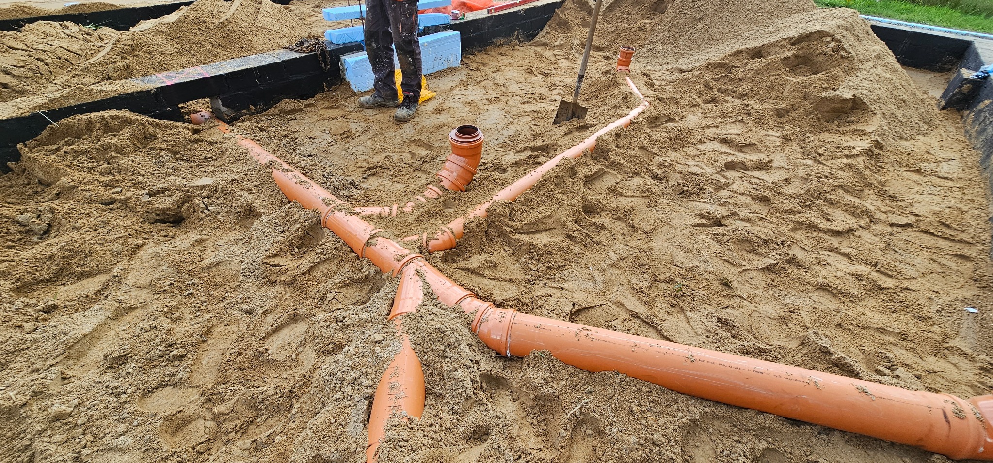 Pomarańczowe rury kanalizacyjne ułożone w piasku na budowie fundamentów, widoczne połączenia trójnikowe i kształtki, w tle pracownik w roboczych butach i fragment izolacji termicznej.