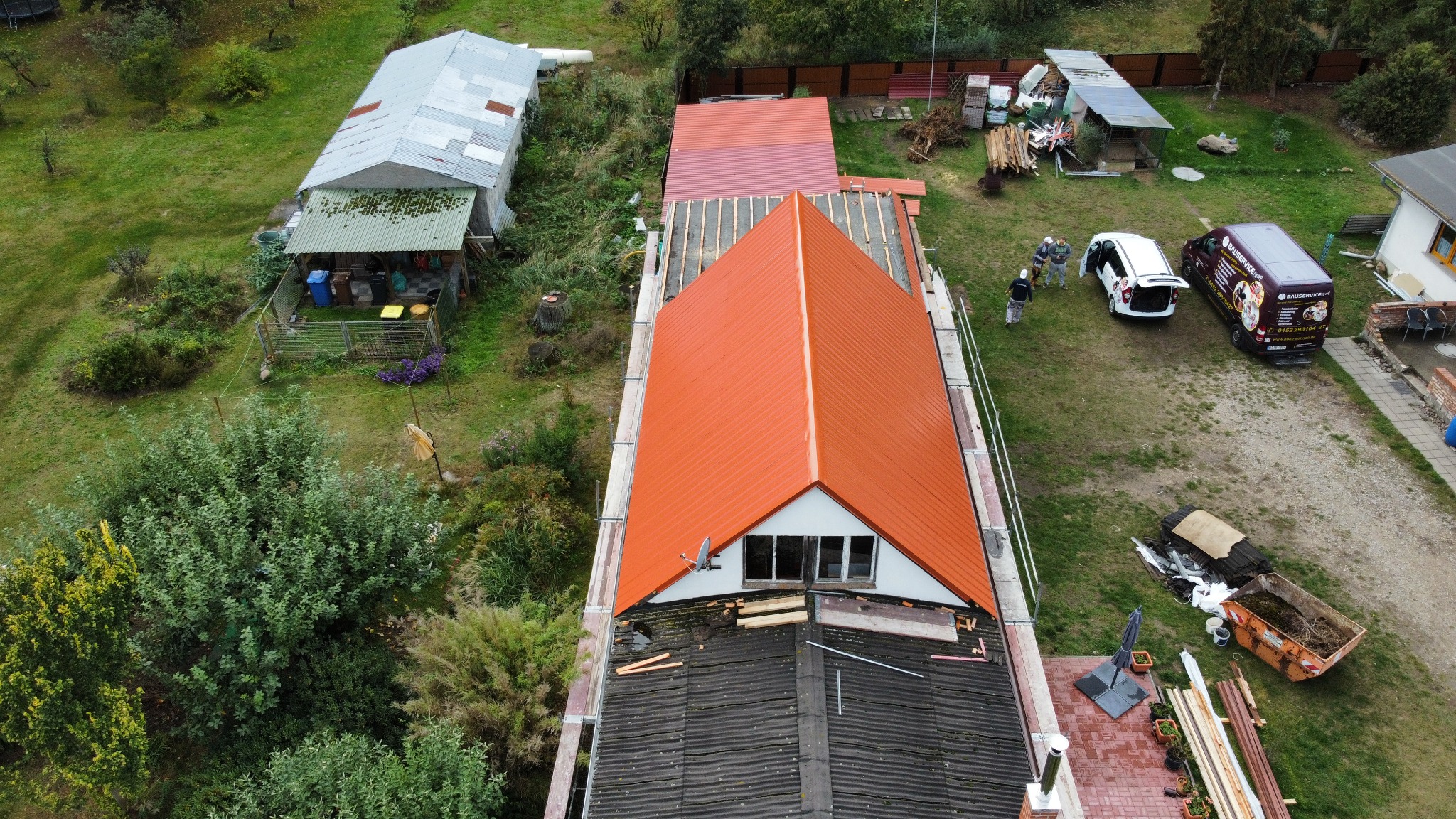 Widok z góry na dom w trakcie wymiany dachu na blachodachówkę w kolorze ceglastym, z widocznymi rusztowaniami, pracownikami i samochodem firmy budowlanej z reklamą na podwórku.