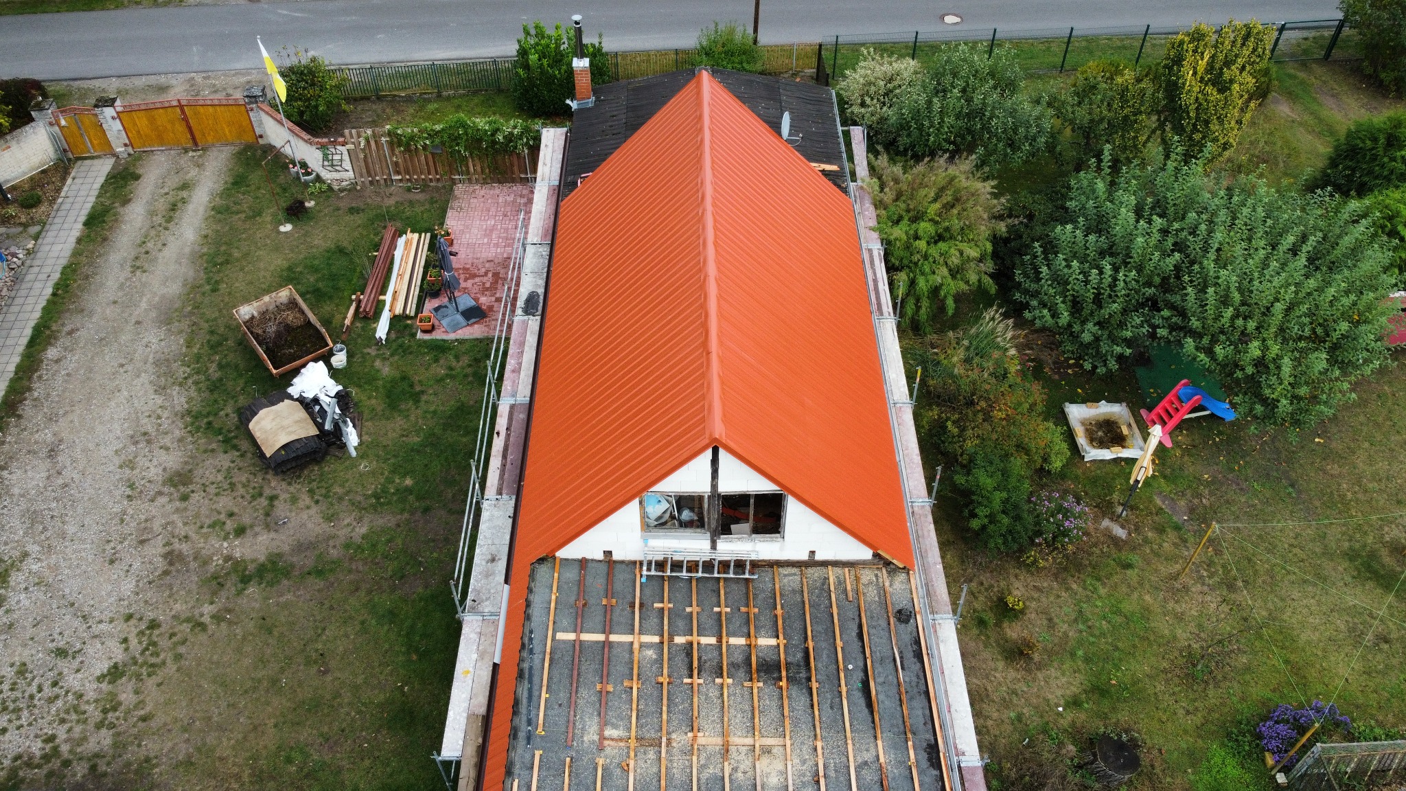 Widok z góry na dom w trakcie wymiany dachu z pomarańczową blachodachówką, widoczne rusztowania i elementy konstrukcji dachu, a w tle ogród z placem zabaw.