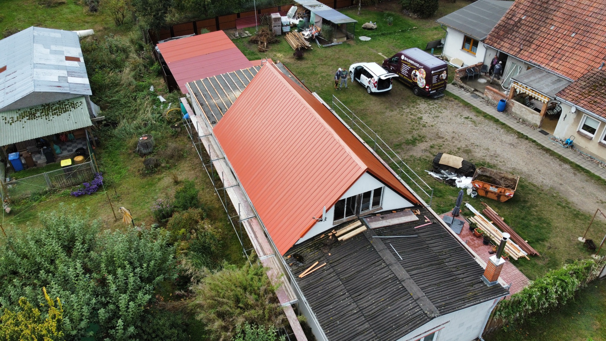 Widok z lotu ptaka na dom w trakcie wymiany dachu z czerwonej blachy, widoczne rusztowania, ekipa remontowa przy samochodzie firmowym i częściowo zdemontowany stary dach.