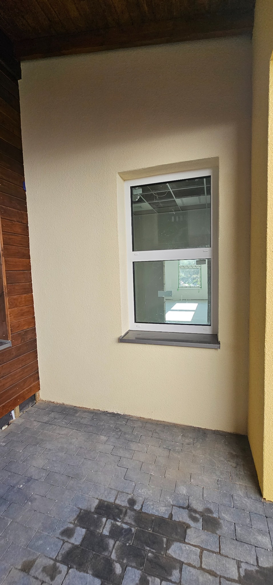 Ściana zewnętrzna budynku po nałożeniu jasnobeżowego tynku strukturalnego, z widocznym oknem z białymi ramami i szarym parapetem oraz fragmentem drewnianej elewacji, położona kostka brukowa.
