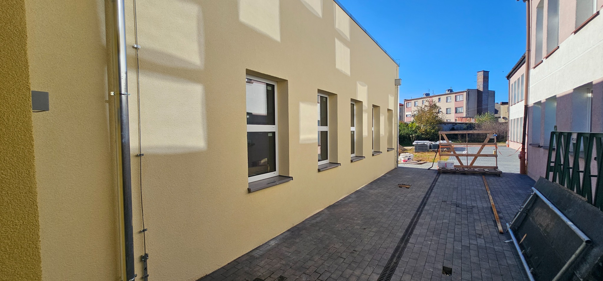 Elewacja budynku po renowacji, z widoczną nową, jasną, teksturowaną farbą, rynnami i nowymi oknami. Na chodniku wzdłuż budynku widoczne materiały budowlane i rusztowanie.