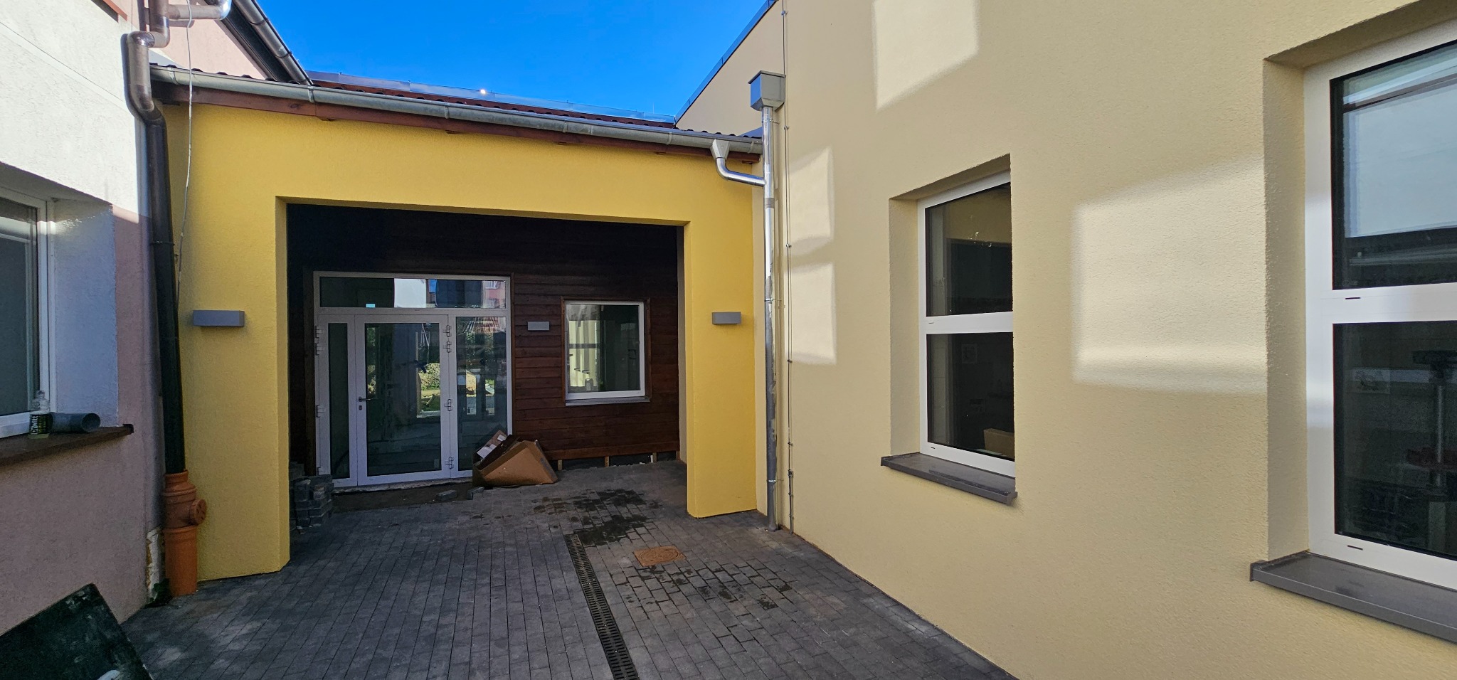 Żółta elewacja budynku z nowymi oknami i wykończonym w drewnie przejściem do posesji, widoczna instalacja odprowadzająca wodę deszczową.