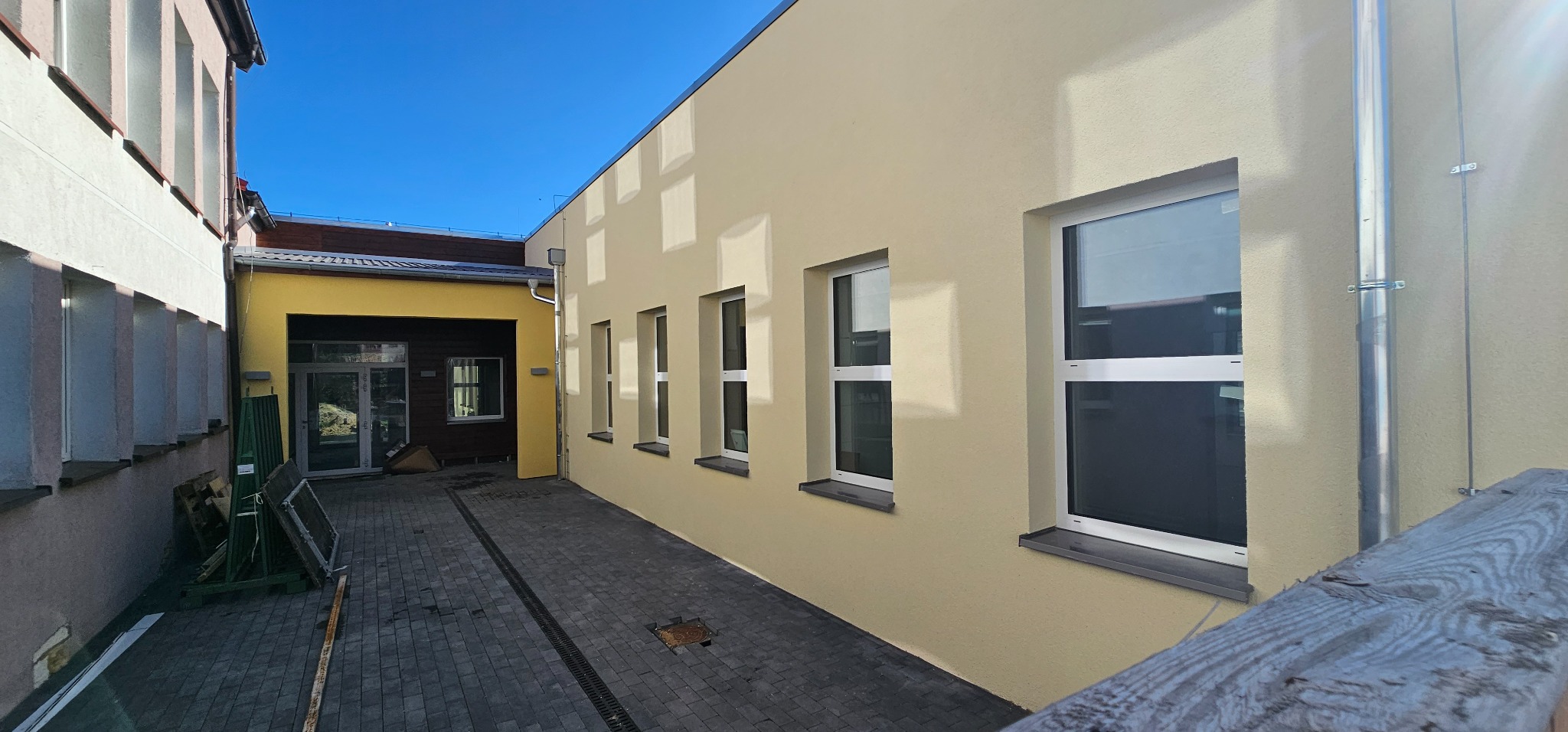 Elewacja budynku po nałożeniu jasnej, beżowej struktury, widoczne okna z białymi ramami i szarymi parapetami, obok zaplecze budowlane i kostka brukowa.