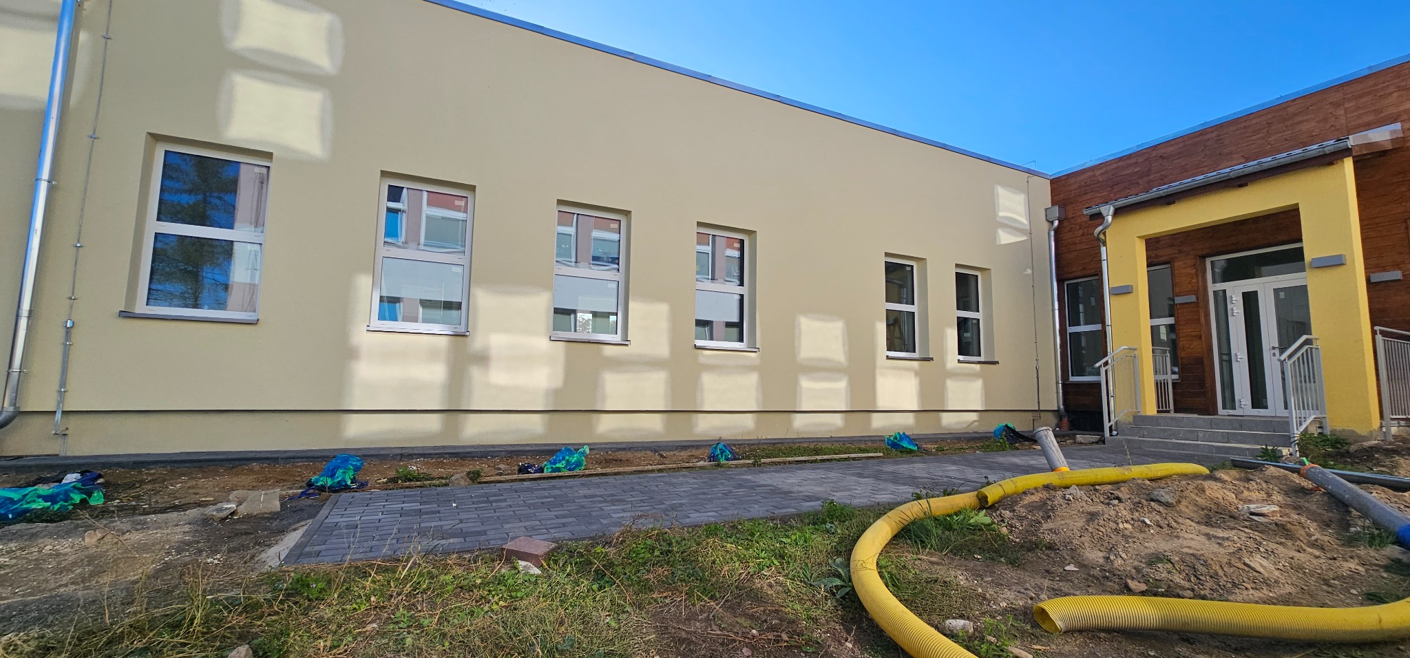 Elewacja budynku po malowaniu na jasny kremowy kolor, widoczne okna z białymi ramami, obok fragment budynku z drewnianą elewacją i żółtym obramowaniem wejścia, teren wokół budynku w trakcie prac...