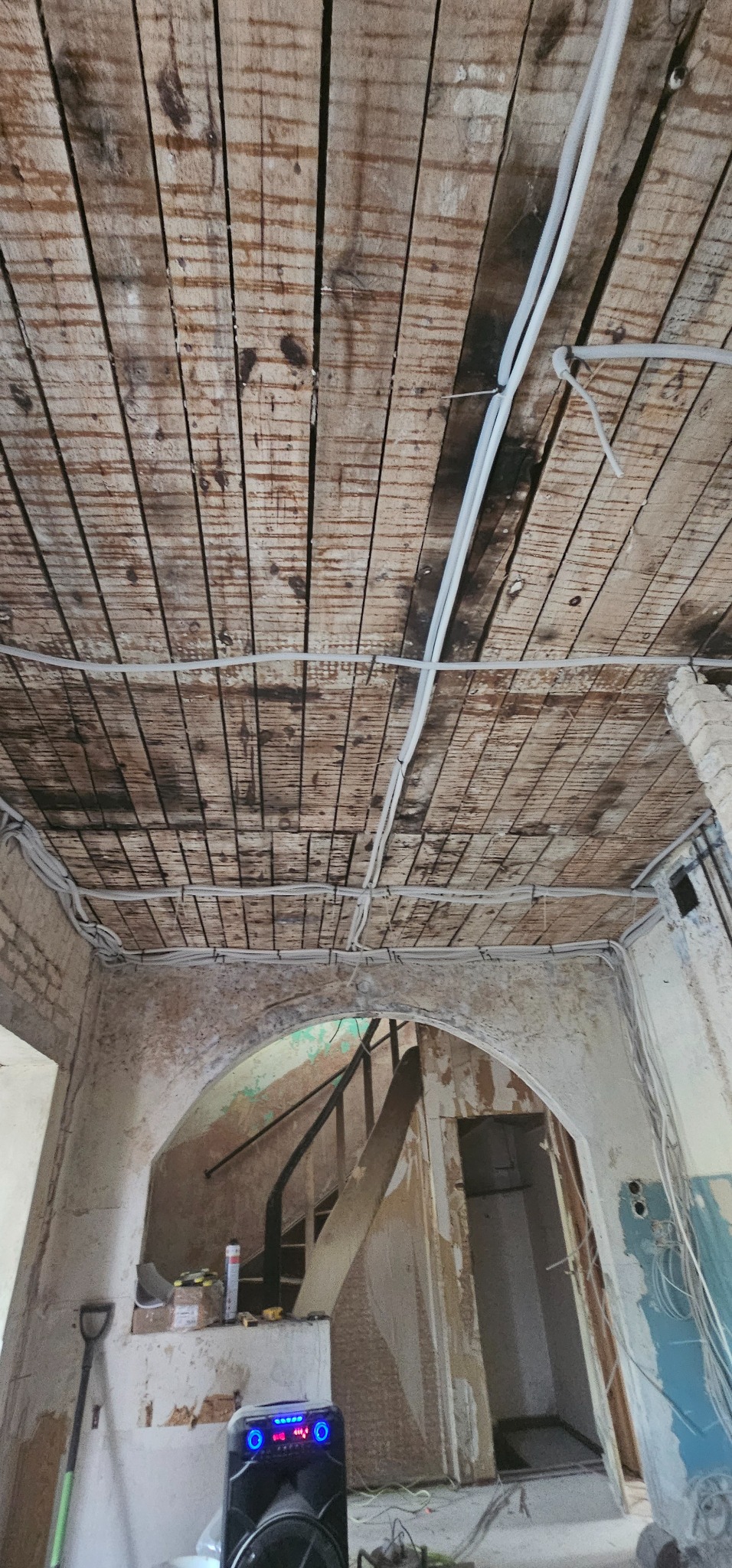 Widok sufitu z drewnianych desek i instalacji elektrycznej w trakcie prac przygotowawczych do remontu, widoczne elementy konstrukcyjne, kable oraz fragment wnętrza z łukowym przejściem i schodami.