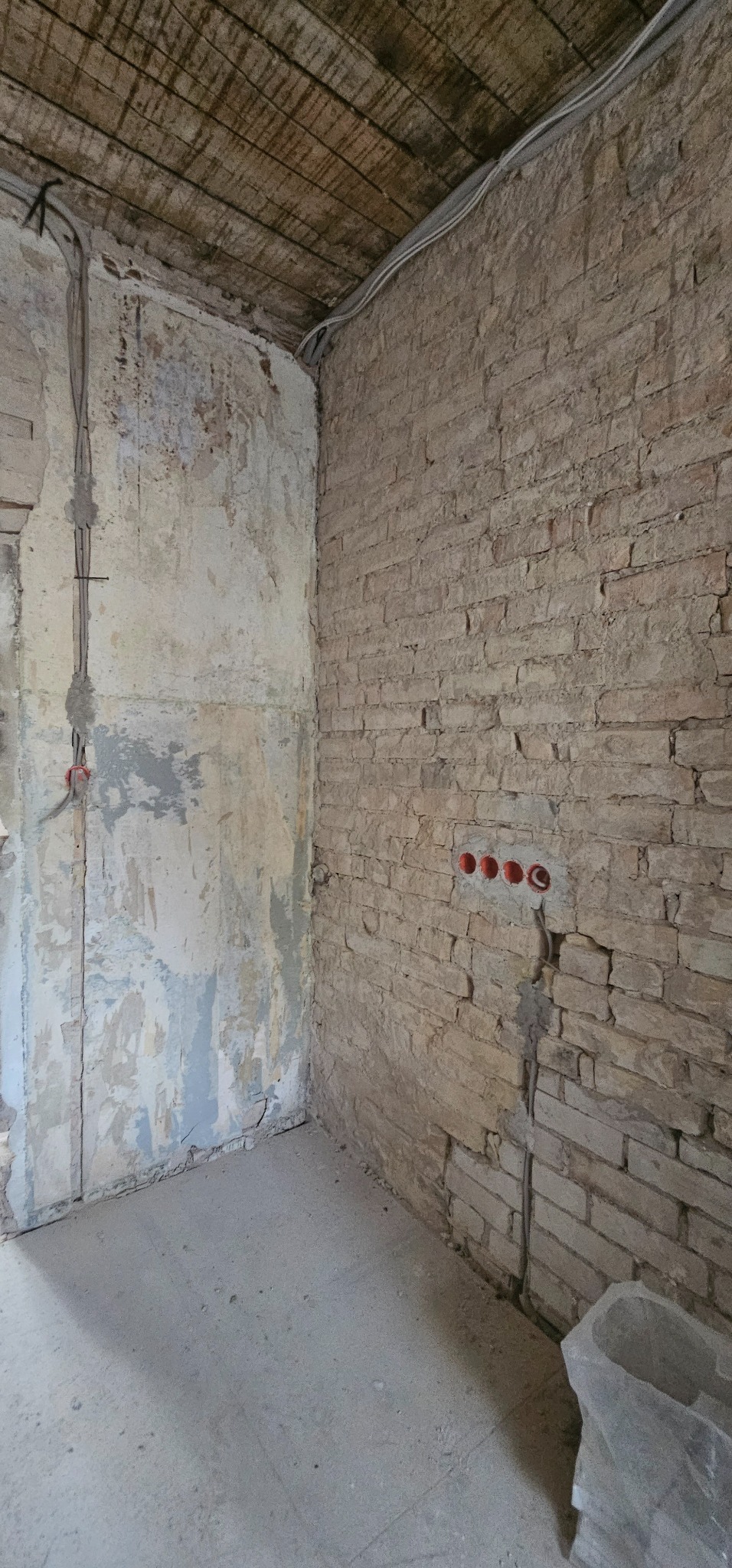 Wnętrze pokoju w trakcie remontu: surowa ceglana ściana z instalacją elektryczną, otynkowana ściana z widocznymi śladami farby, odsłonięty drewniany strop i betonowa podłoga.