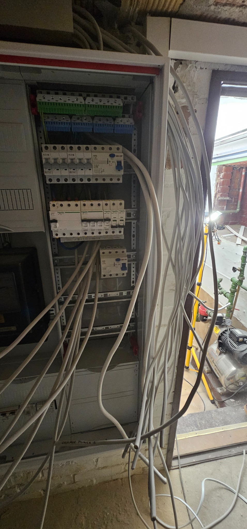 Otwarta skrzynka elektryczna z bezpiecznikami i licznymi przewodami, widoczne również fragmenty ściany z cegły i pompy hydroforowej w tle.