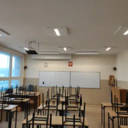 Sala lekcyjna z rzędami ławek z krzesłami na nich, biała tablica, wiszący projektor i panele oświetleniowe LED na suficie.