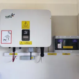 Biała obudowa inwertera fotowoltaicznego marki Selfa z widocznymi naklejkami ostrzegawczymi, podłączona do przewodów i rozdzielnicy elektrycznej na białej ścianie. Widoczny fragment instalacji...