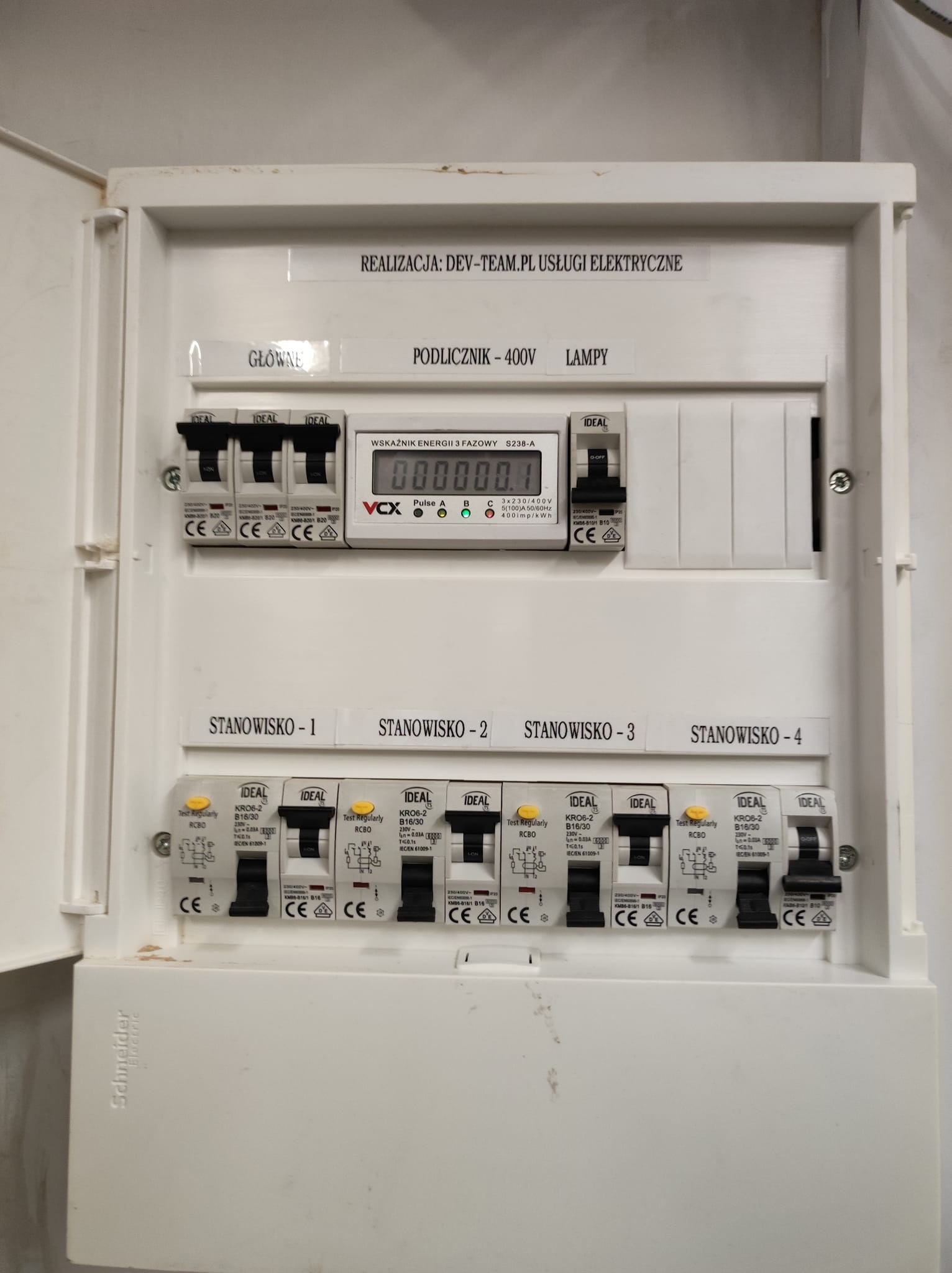 Otwarta, biała skrzynka rozdzielcza z bezpiecznikami i licznikiem energii, oznaczona jako 'Stanowisko - 1' do 'Stanowisko - 4', z naklejką 'REALIZACJA: DEV-TEAM.PL USŁUGI ELEKTRYCZNE'.