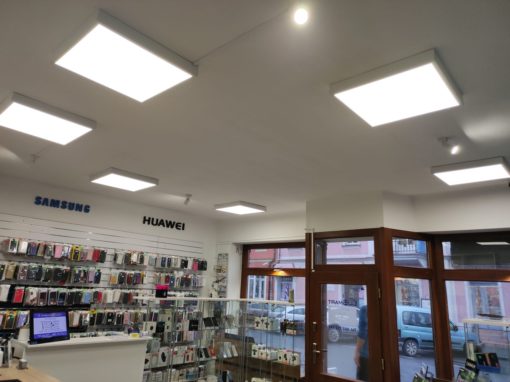 Sklep z akcesoriami GSM oświetlony panelami LED, widok na sufit z sześcioma kwadratowymi lampami i ekspozycję etui na telefony marek Samsung i Huawei w tle.