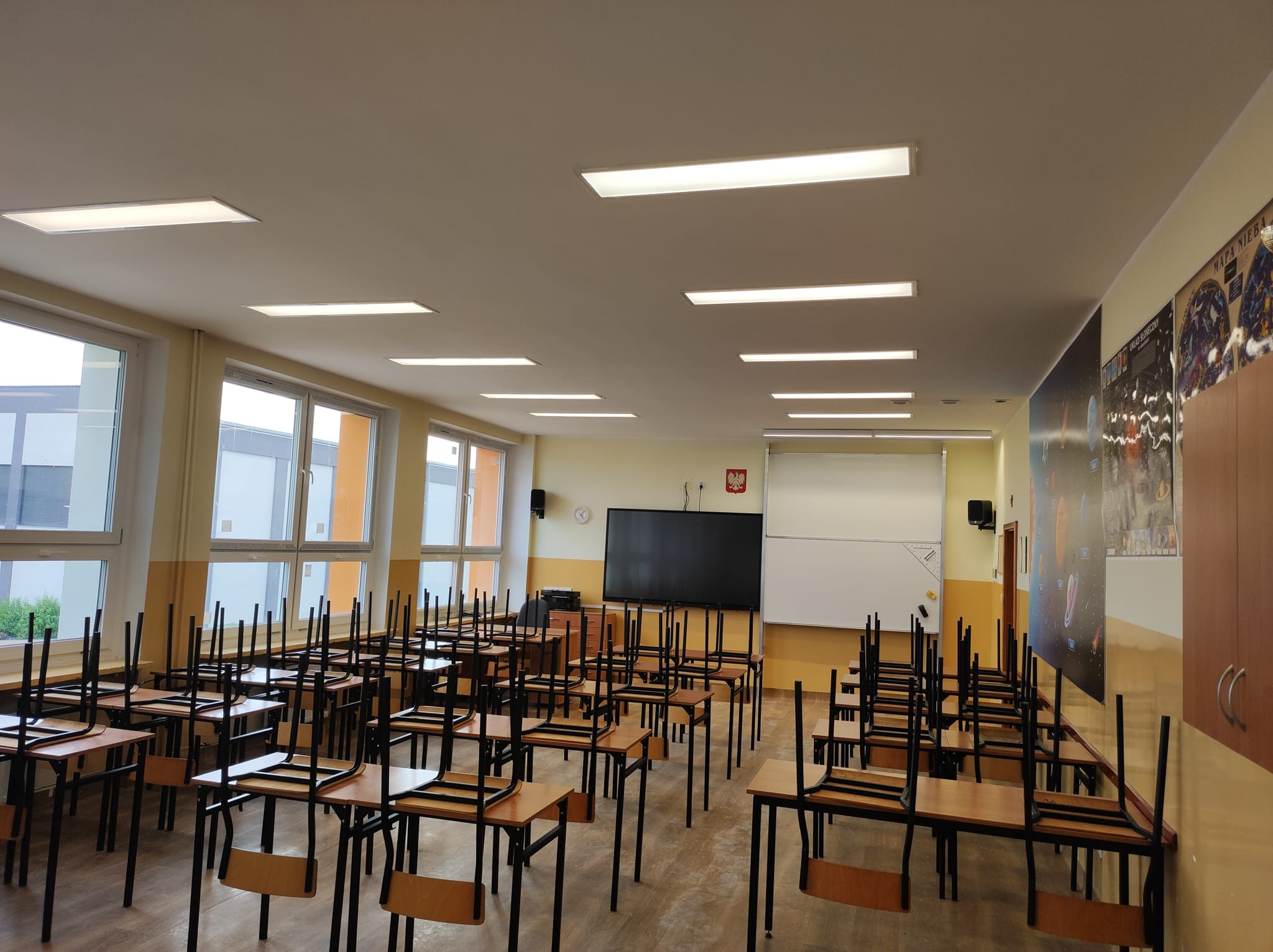 Wnętrze pustej sali lekcyjnej z rzędami ławek i krzeseł postawionymi na blatach, oświetlonej panelami LED w suficie.