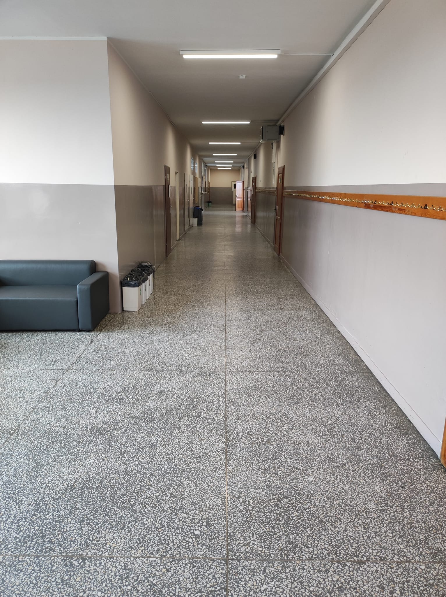 Długi korytarz w budynku użyteczności publicznej z szarym lastryko na podłodze, białymi ścianami z szarym pasem, rzędem drzwi po obu stronach, oświetleniem jarzeniowym na suficie, sofą i koszami...