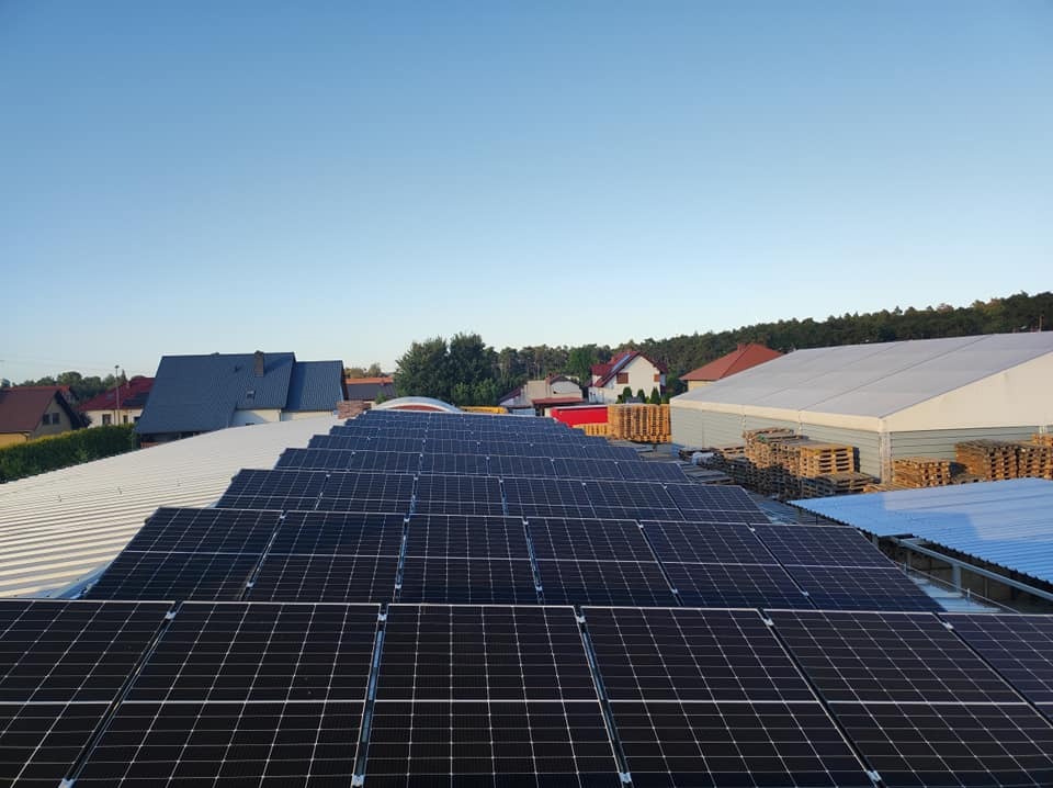 Rząd paneli fotowoltaicznych na dachu hali magazynowej, w tle domy jednorodzinne i błękitne niebo.