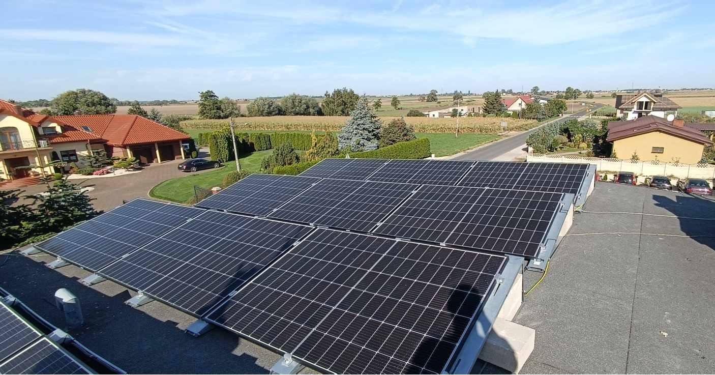 Panele fotowoltaiczne na dachu domu jednorodzinnego, widok z góry na okolicę z zabudową mieszkalną i polami uprawnymi w słoneczny dzień.