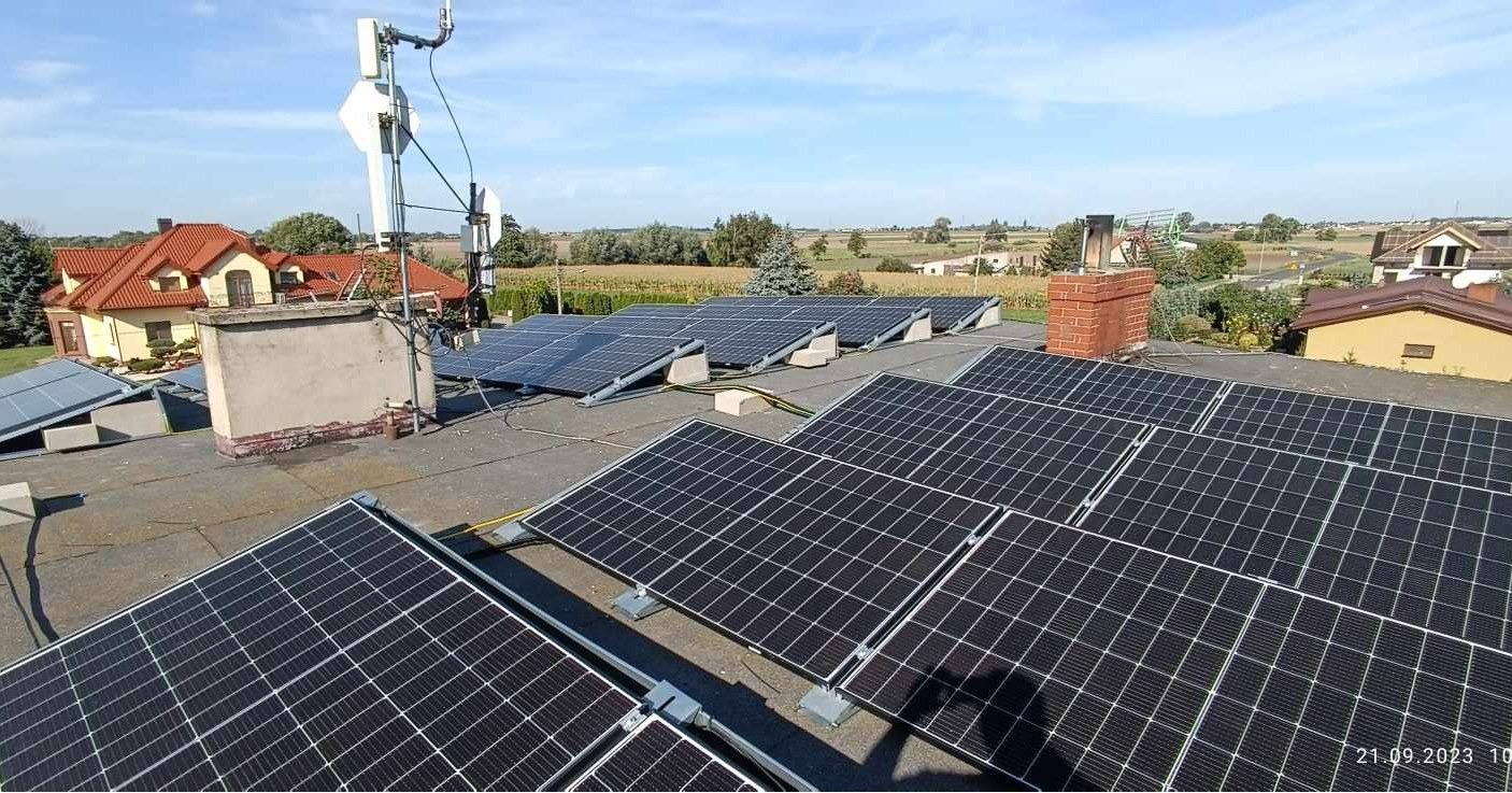 Widok z góry na panele fotowoltaiczne zamontowane na płaskim dachu budynku, z widocznym kominem z cegły i anteną telekomunikacyjną w tle krajobrazu wiejskiego pod błękitnym niebem.