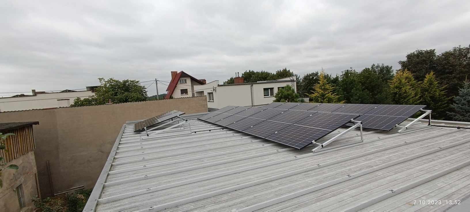 Panele fotowoltaiczne zamontowane na metalowym dachu z widokiem na budynki mieszkalne i drzewa w tle, pochmurne niebo.