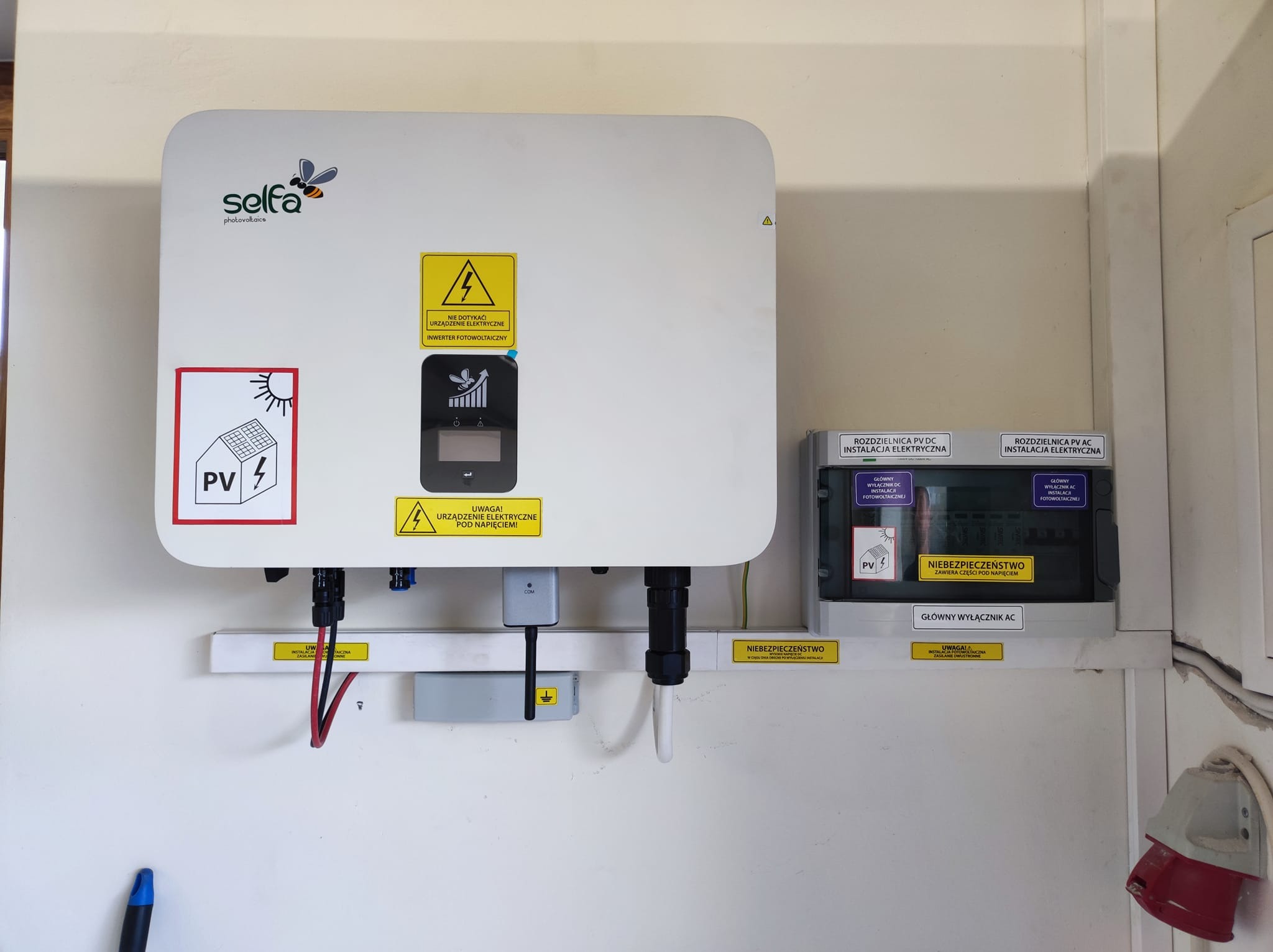 Biała obudowa inwertera fotowoltaicznego marki Selfa z widocznymi naklejkami ostrzegawczymi, podłączona do przewodów i rozdzielnicy elektrycznej na białej ścianie. Widoczny fragment instalacji...