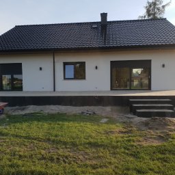 Lew Król Twojego Ogrodu - Nowoczesny dom jednorodzinny z ciemną dachówką i tarasem w trakcie budowy, widoczna taczka i przygotowane płyty tarasowe.