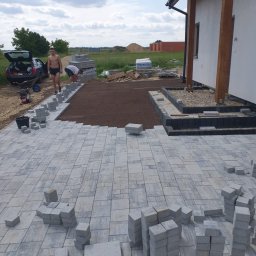 Lew Król Twojego Ogrodu - Prace brukarskie w toku: ułożona część szarej kostki brukowej, przygotowane podłoże z brązowej ziemi, pracownicy w trakcie układania, widoczny samochód z otwartym bagażnikiem i narzędzia.