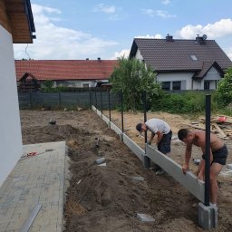 Lew Król Twojego Ogrodu - Dwóch monterów umieszcza betonowe elementy podmurówki ogrodzeniowej na słupkach, widoczne narzędzia i materiały budowlane, w tle domy jednorodzinne.