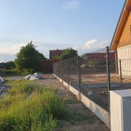 Lew Król Twojego Ogrodu - Nowo postawione ogrodzenie panelowe z podmurówką betonową, widoczne worki z materiałami budowlanymi w tle, wysoka trawa z przodu, dom z cegły i drewniany dom w oddali.