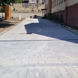 Świeżo ułożona, szara kostka brukowa na placu budowy w słoneczny dzień, z pracownikiem i budynkami w tle.