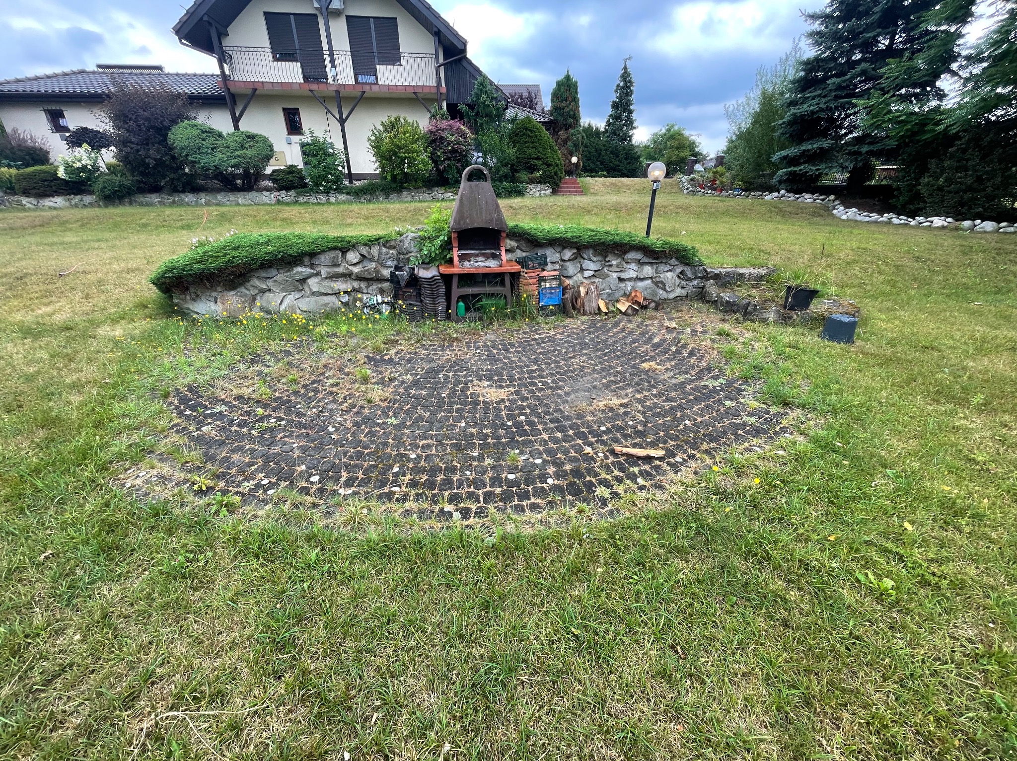 Półokrągła powierzchnia z ażurowych płyt betonowych na trawniku, otoczona niskim kamiennym murkiem z grillem, drewnem i narzędziami, na tle domu z ogrodem.