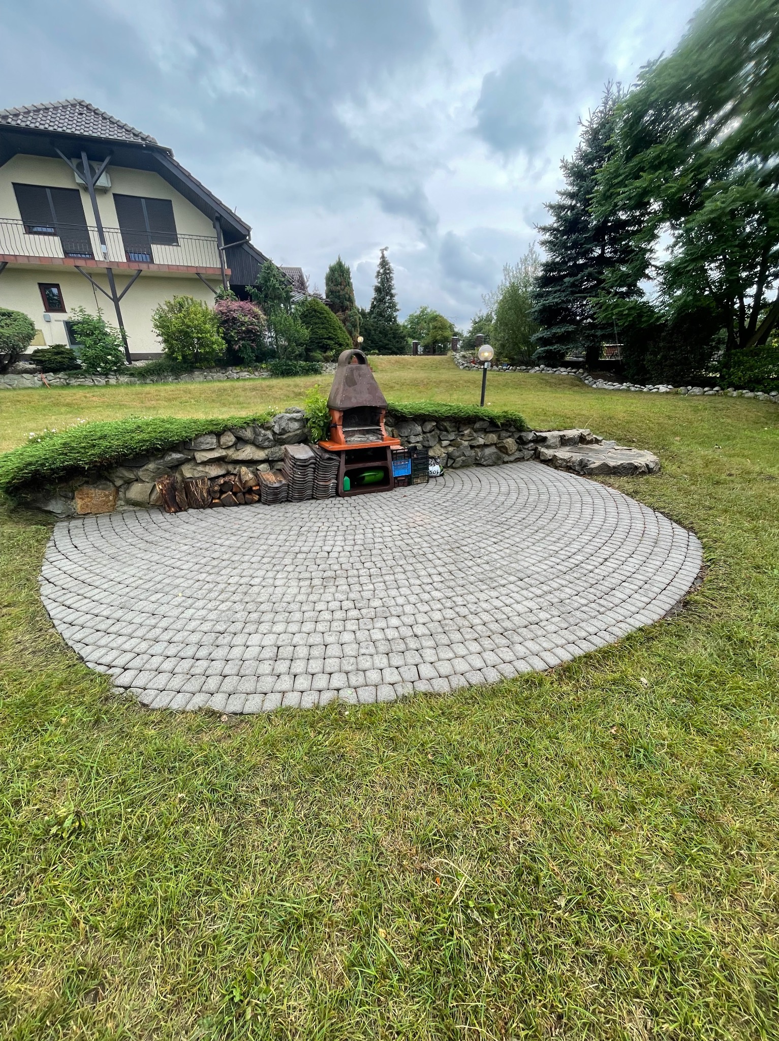 Półokrągły taras z kostki brukowej w ogrodzie z grillem murowanym, ułożony z szarej kostki w okrągły wzór, na tle domu i drzew.