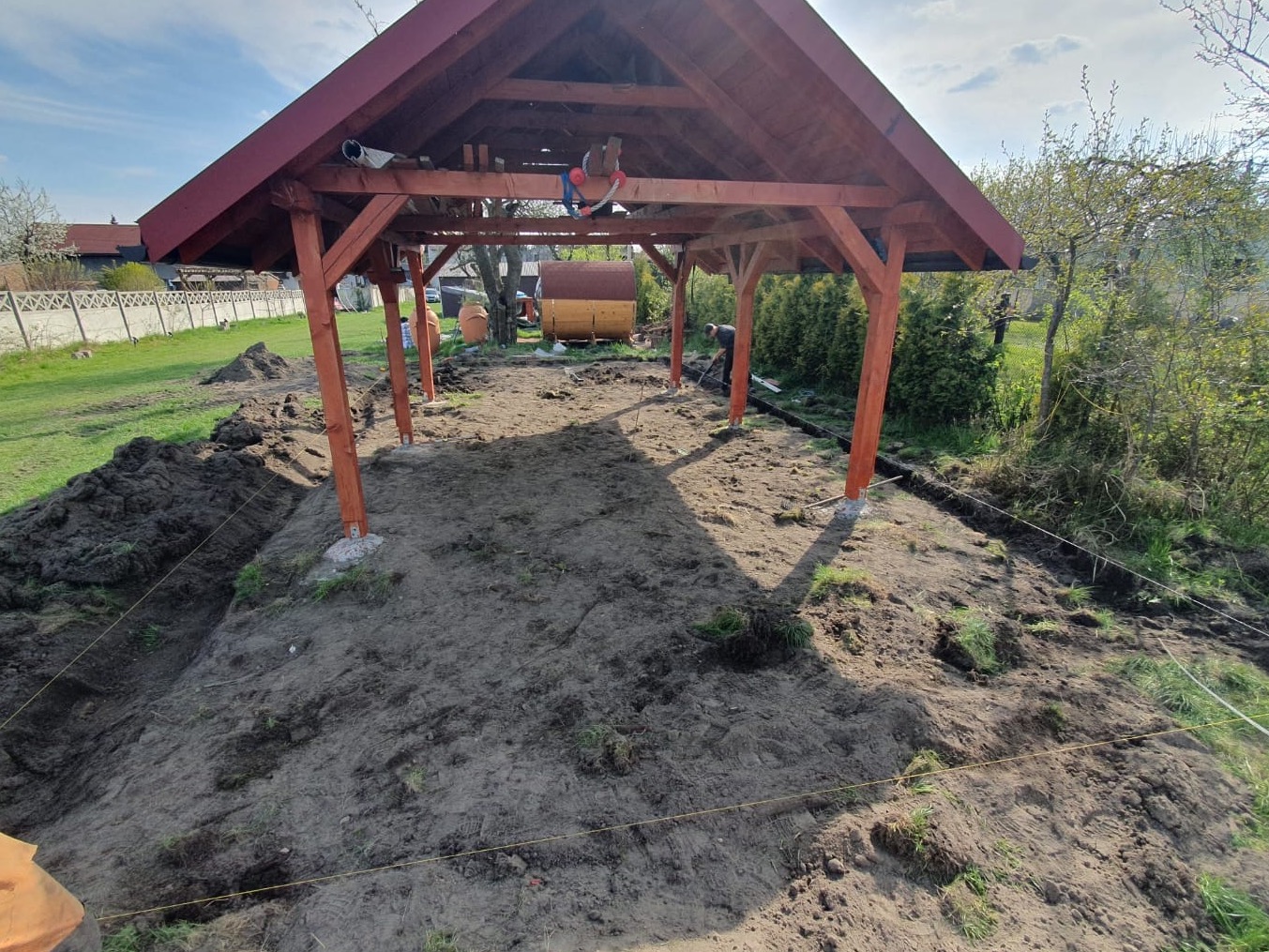 Konstrukcja drewnianej altany w trakcie budowy, widoczne słupy z betonowymi podstawami, przygotowany teren i pracownik wyrównujący ziemię łopatą.