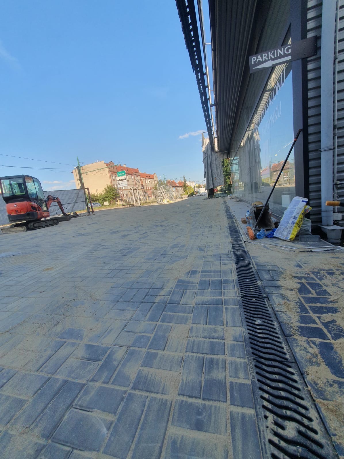 Nowo ułożona, szara kostka brukowa na chodniku przed budynkiem z szyldem 'PARKING', w tle mini koparka i materiały budowlane.