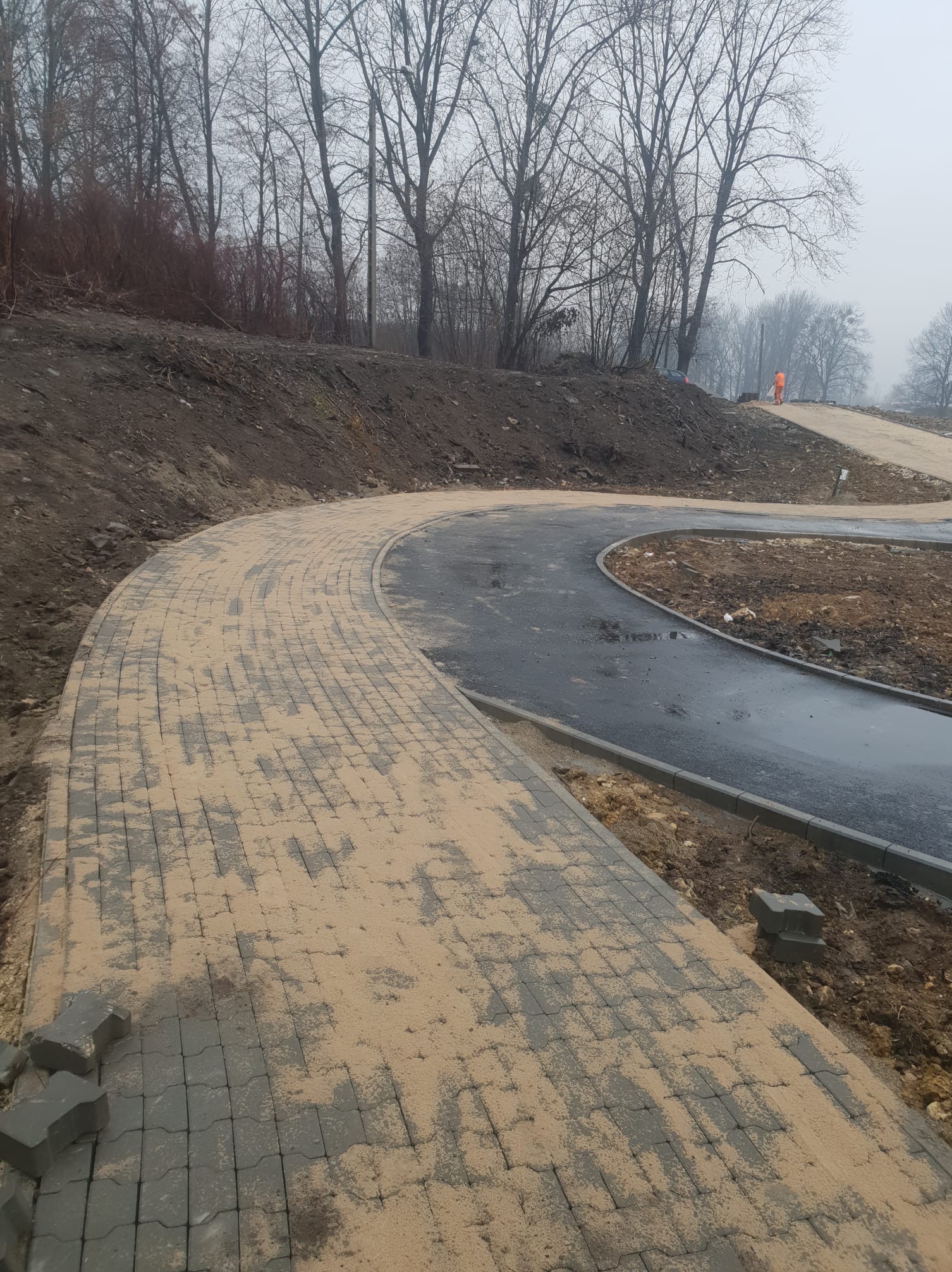 Układanie zakrzywionego chodnika z szarej kostki brukowej, częściowo zasypanej piaskiem, z widocznym fragmentem asfaltowej ścieżki i pracownikiem w oddali w pomarańczowym kombinezonie.