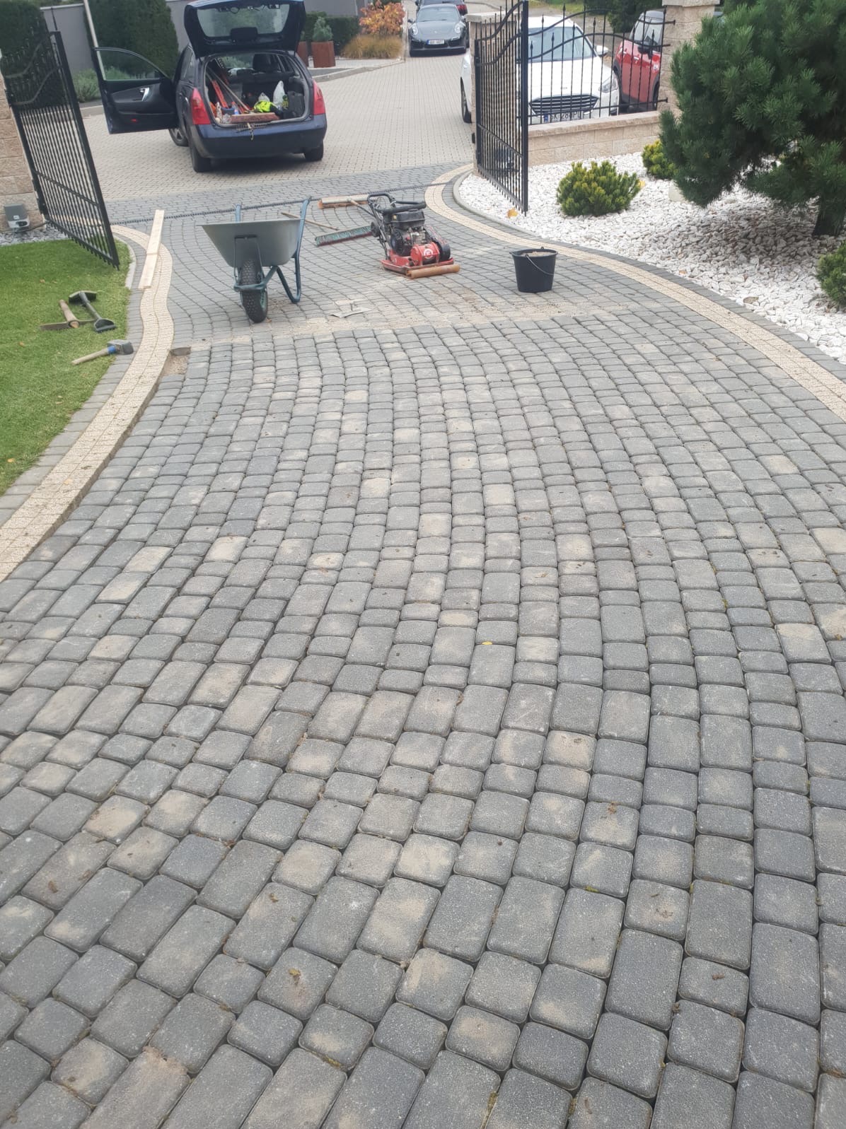 Ułożona, szara kostka brukowa na podjeździe, widoczna ubijarka, taczka i narzędzia, samochód z otwartym bagażnikiem w tle.