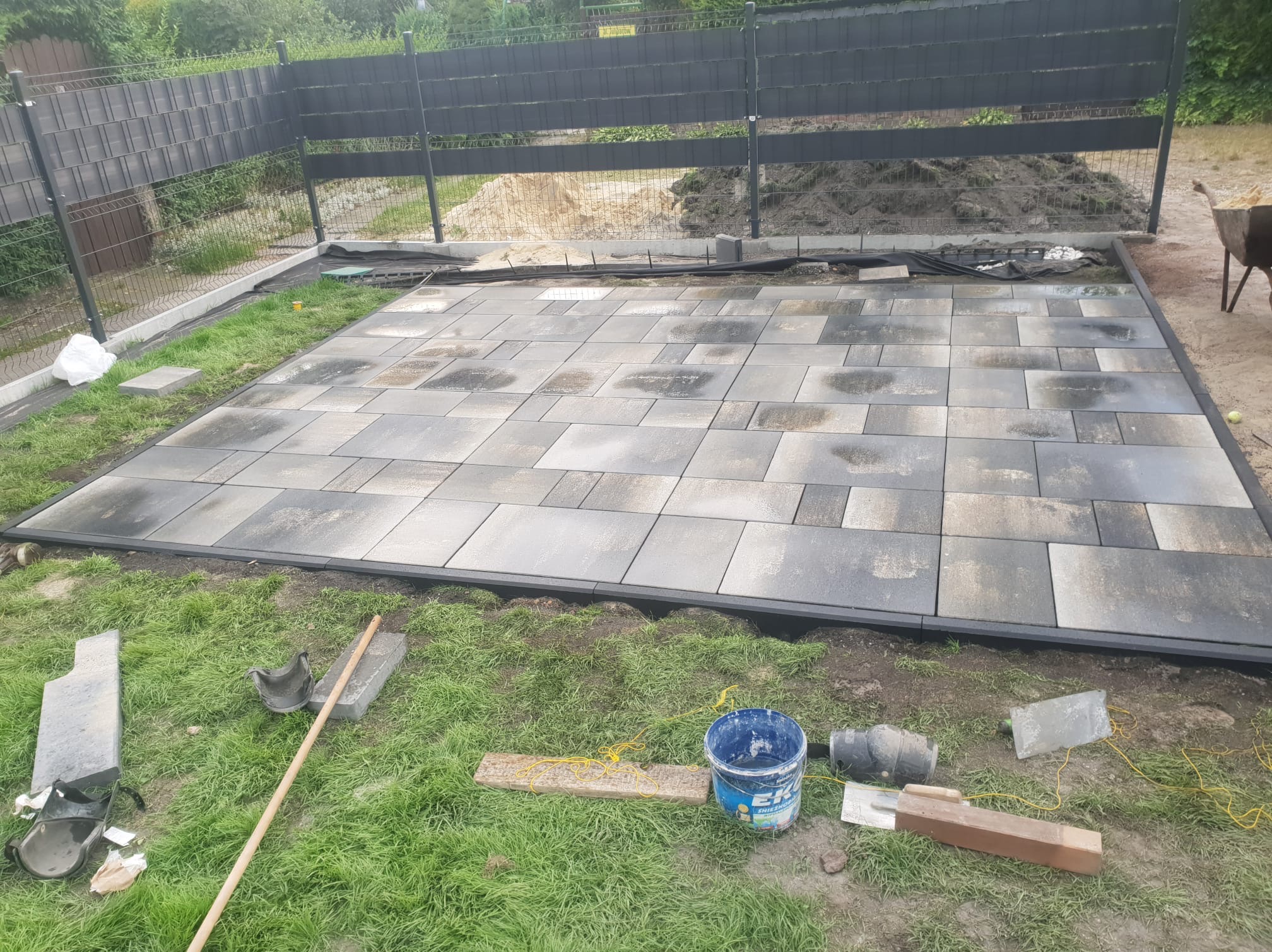 Częściowo ułożona, prostokątna nawierzchnia z kostki brukowej w Zabrzu, obok narzędzia i wiadro, w tle ogrodzenie panelowe.