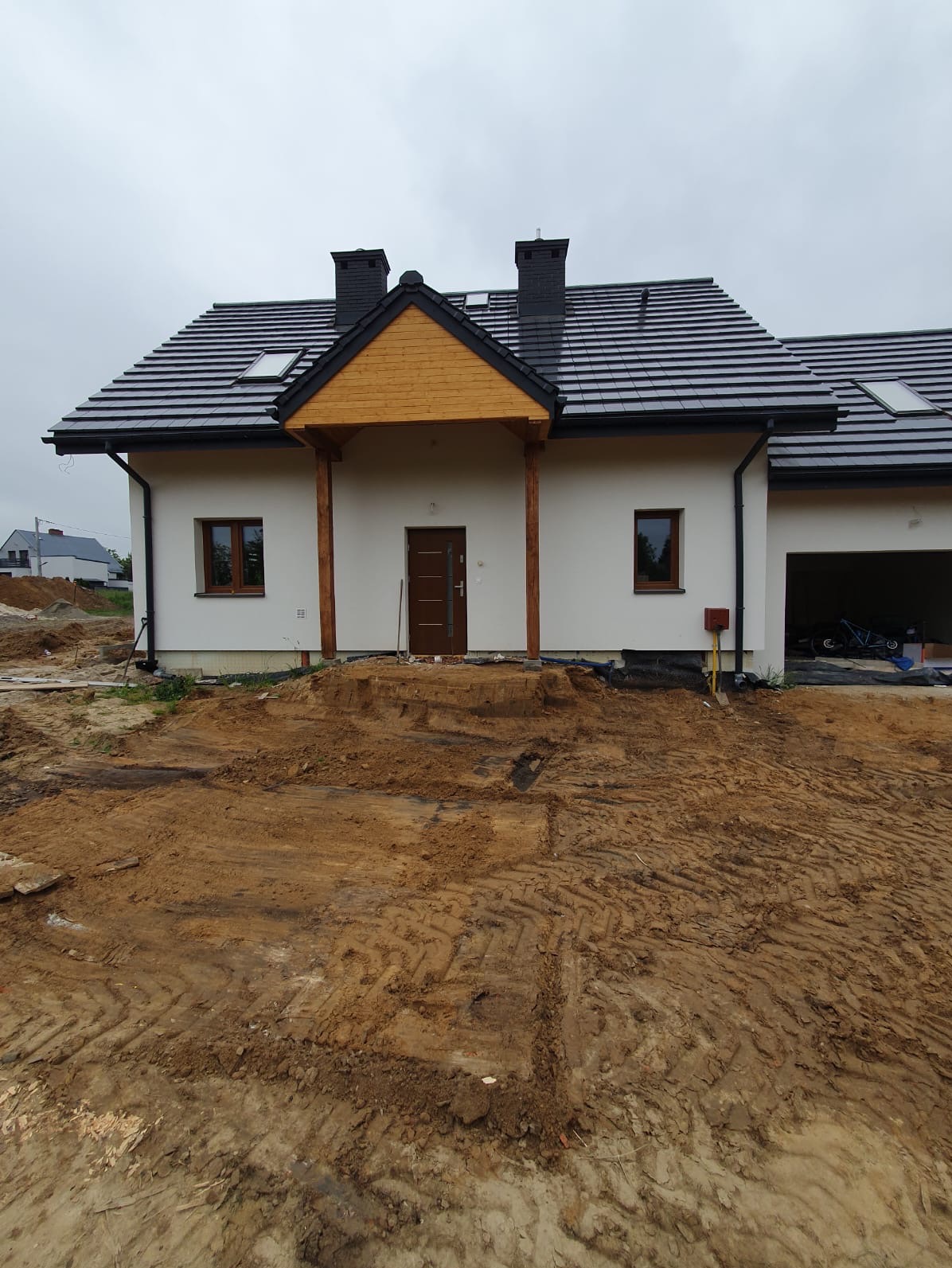 Nowoczesny dom jednorodzinny z białymi ścianami, drewnianą wiatą nad wejściem i czarną dachówką, otoczony rozkopanym terenem przygotowanym pod zagospodarowanie, widok na front budynku.