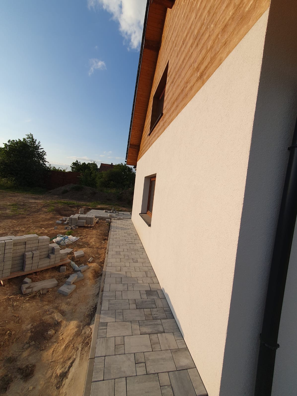 Świeżo ułożona, szara kostka brukowa tworzy chodnik wzdłuż ściany domu z drewnianą górną częścią. Stosy kostki czekają na ułożenie. Widoczne okno w ścianie budynku.