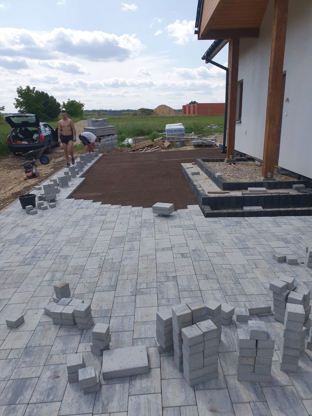 Prace brukarskie w toku: ułożona część szarej kostki brukowej, przygotowane podłoże z brązowej ziemi, pracownicy w trakcie układania, widoczny samochód z otwartym bagażnikiem i narzędzia.