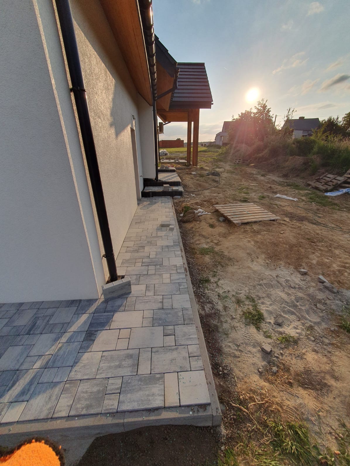 Ułożona kostka brukowa wzdłuż boku domu z widoczną rynną spustową, w tle niezagospodarowany teren z paletami i zachodzące słońce.