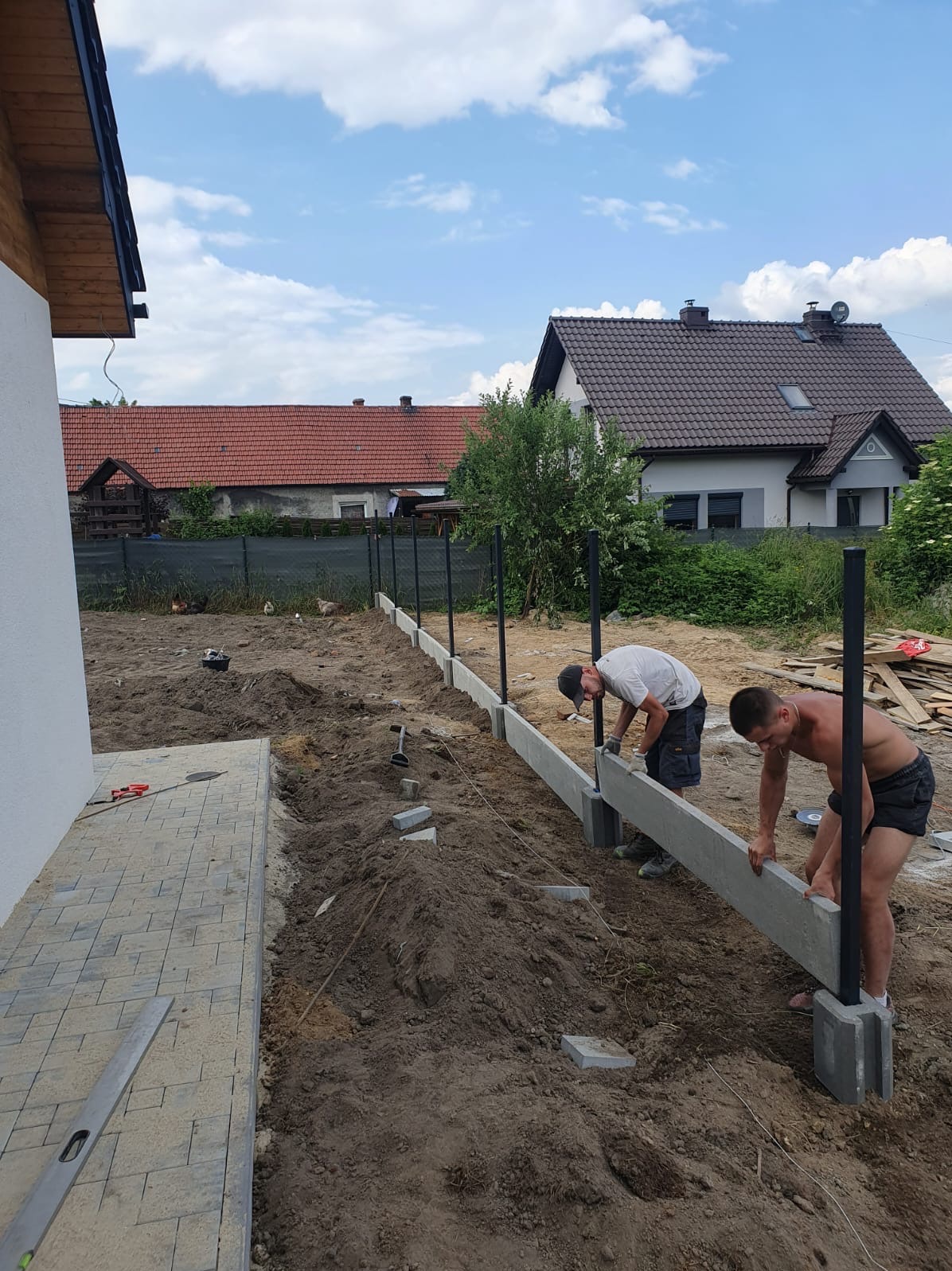Dwóch monterów umieszcza betonowe elementy podmurówki ogrodzeniowej na słupkach, widoczne narzędzia i materiały budowlane, w tle domy jednorodzinne.